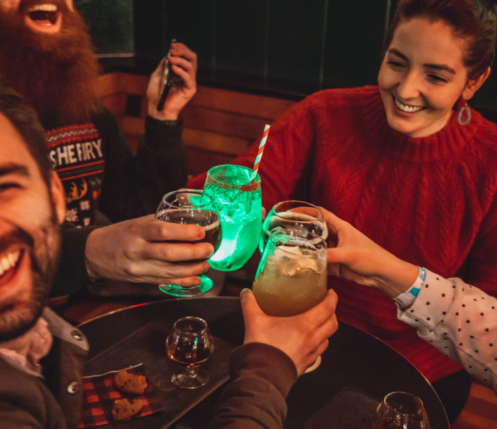 Four people clink glasses at a cozy bar. They hold a variety of drinks, including one with a glowing green light. The group is smiling and wearing casual winter clothing, exuding a warm and festive atmosphere.